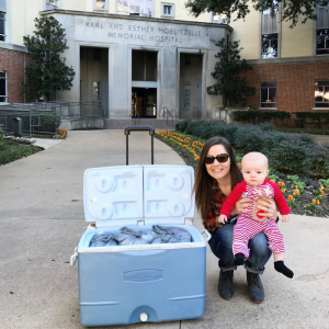 Mother holding baby, crouching next to cooler of frozen breastmilk